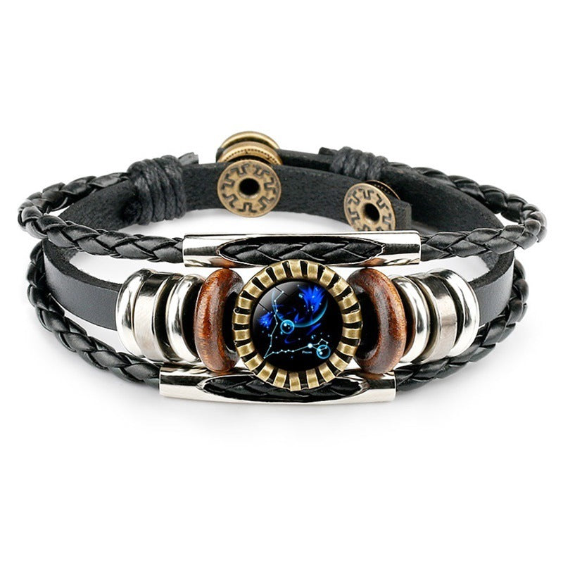 A stylish black leather bracelet with braided straps, featuring metal and wooden beads. The centerpiece is a round, artful design displaying a vivid blue pattern.