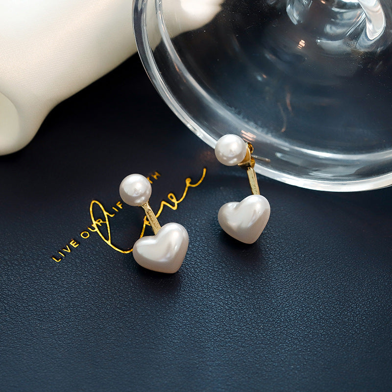 Pearl heart-shaped earrings on a dark surface with a clear container and white object in the background.