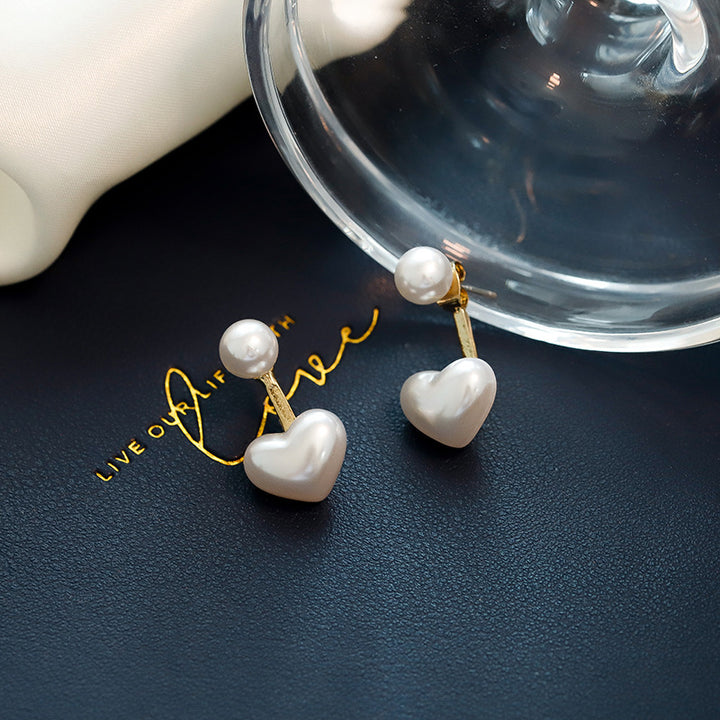 Pearl heart-shaped earrings on a dark surface with a clear container and white object in the background.