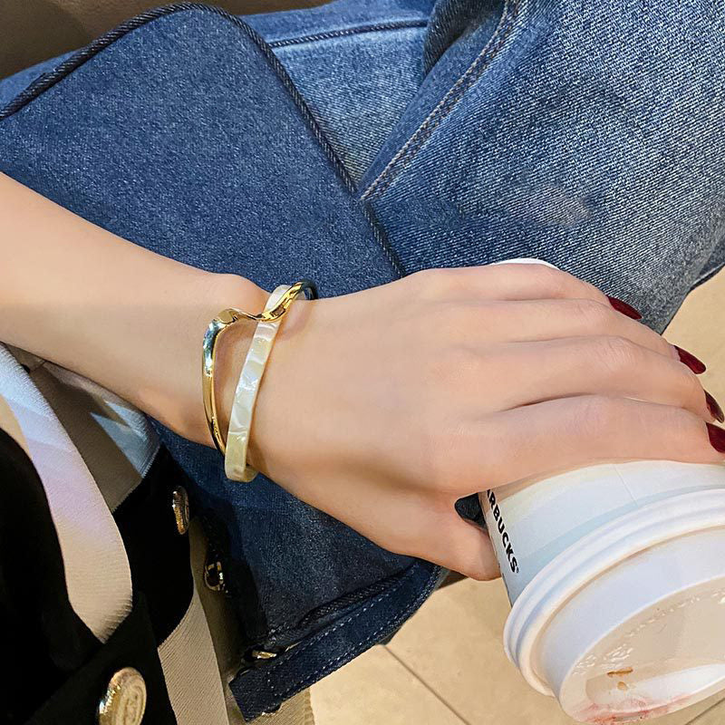 Person wearing gold bracelets holding a Starbucks cup with a denim jacket.