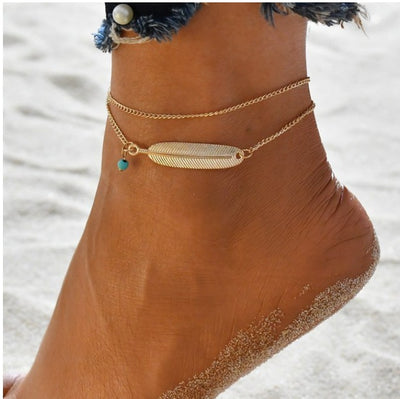 Gold anklet with a feather charm on a person's ankle against a sandy background