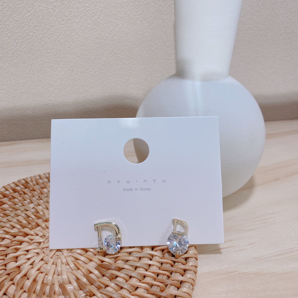 Gold earrings with clear stones on a white card with 'nyonya' branding, placed on a woven mat.