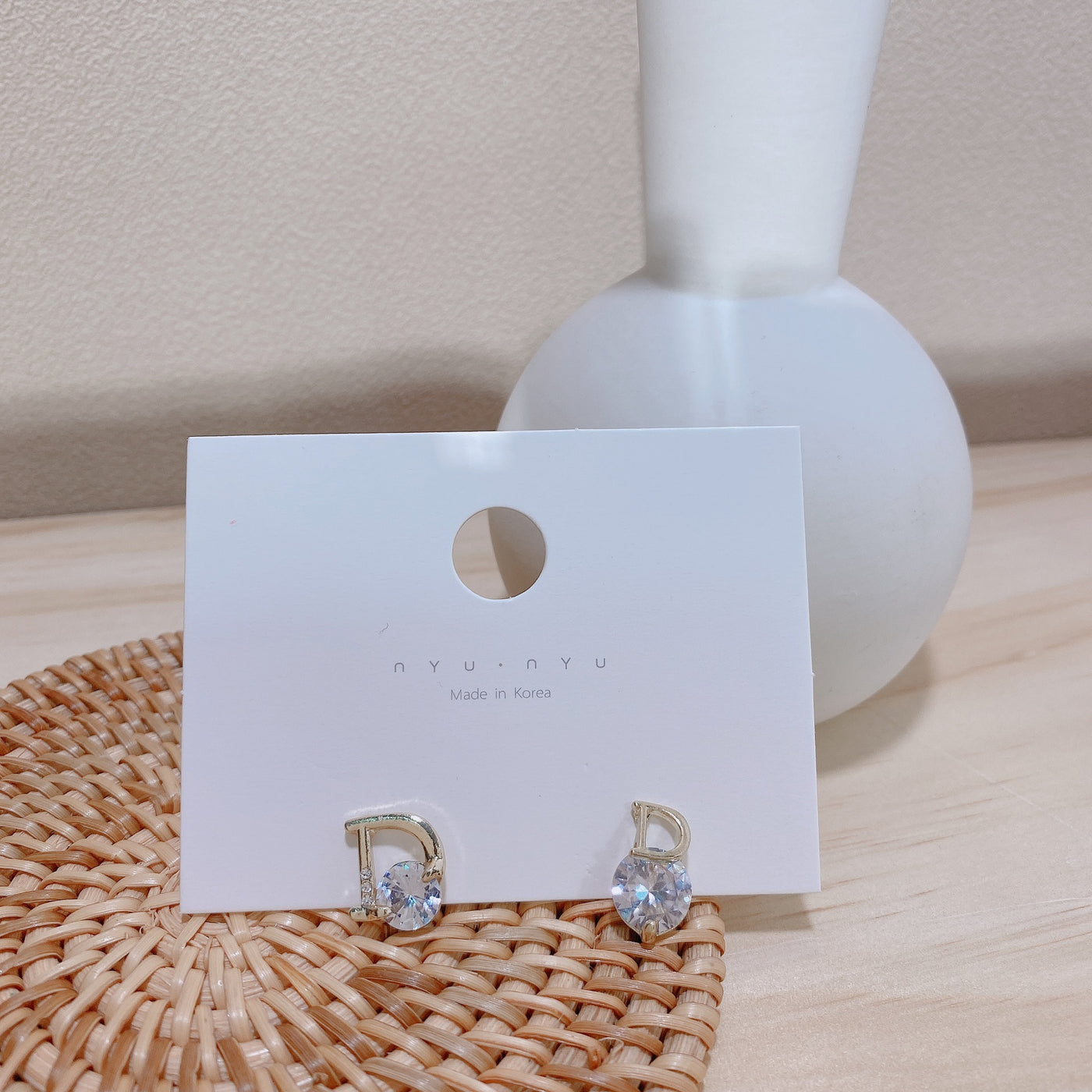 Gold earrings with clear stones on a white card with 'nyonya' branding, placed on a woven mat.