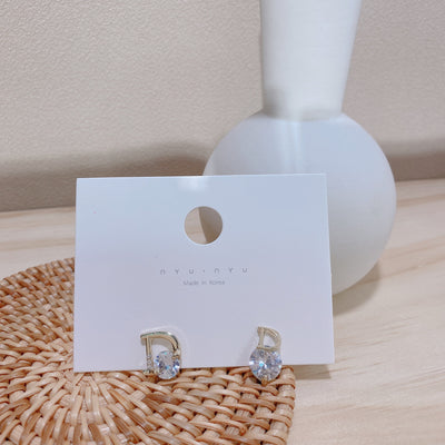 Gold earrings with clear stones on a white card with 'nyonya' branding, placed on a woven mat.