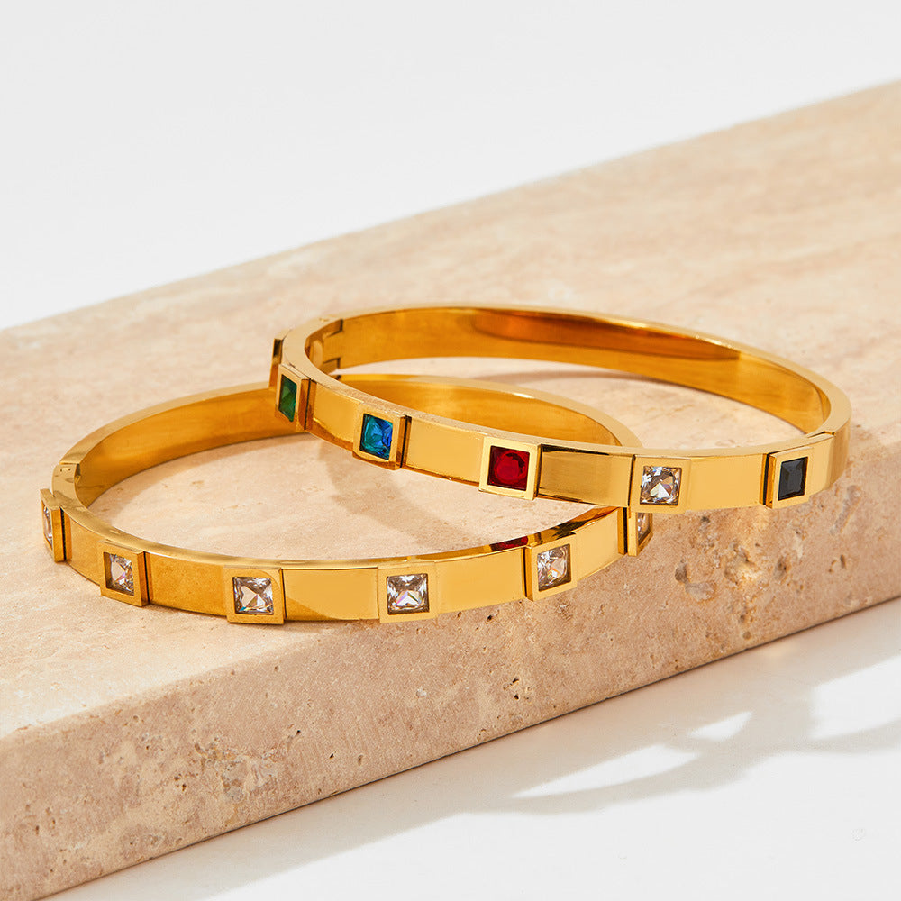 Two gold bangles with embedded gemstones on a beige stone surface
