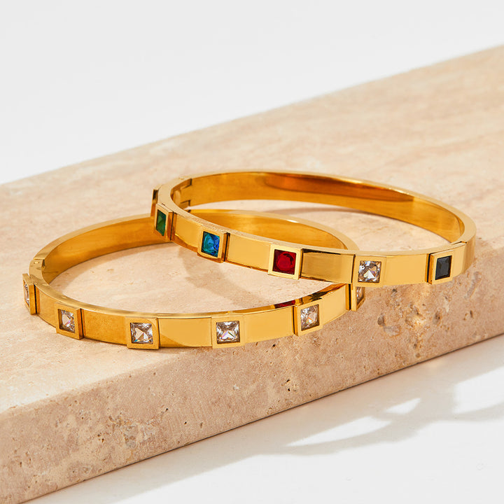 Two gold bangles with embedded gemstones on a beige stone surface