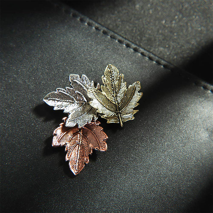Three leaf-shaped pins in silver, gold, and rose gold on a dark surface