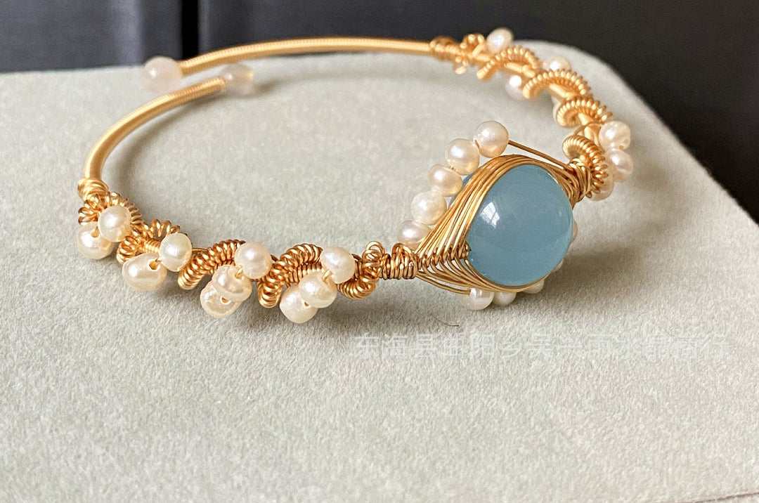 left side view of a Gold bracelet with pearls and a blue stone on a light gray surface