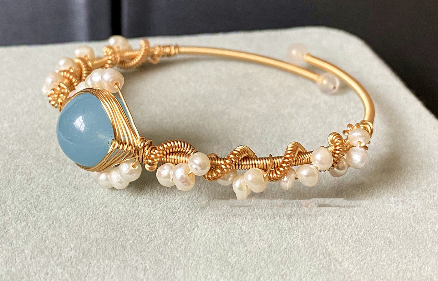 side view of a Gold bracelet with pearls and a blue stone on a light gray surface