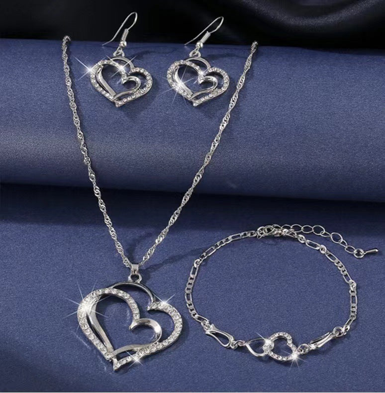 Silver heart-shaped jewelry set including necklace, earrings, and bracelet on a blue fabric background