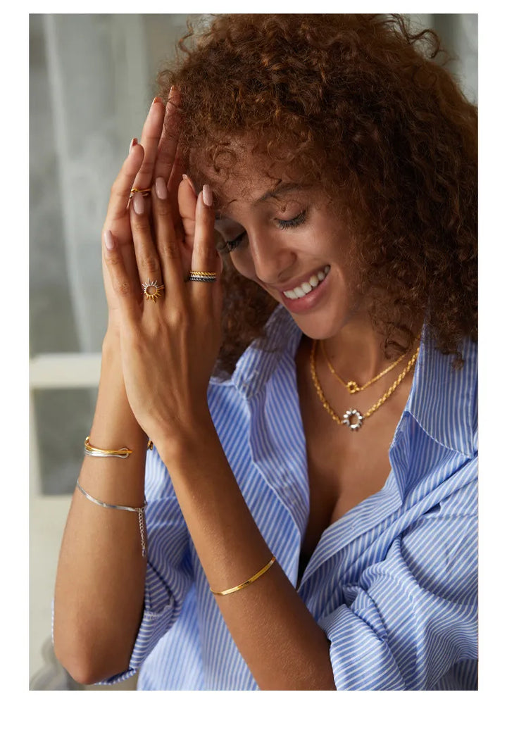 Woman wearing gold jewelry including a necklace, bracelet, and ring, with a blurred background.