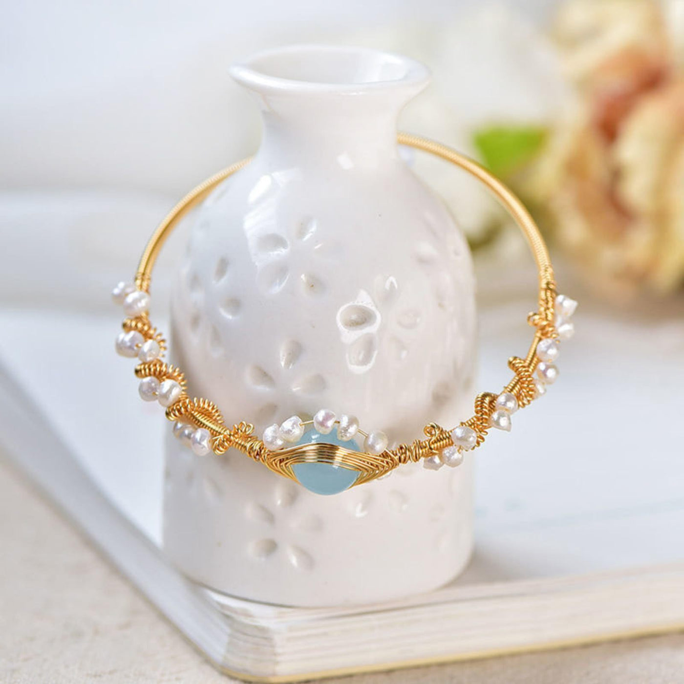 Gold and pearl bracelet on a white ceramic vase with a blurred floral background