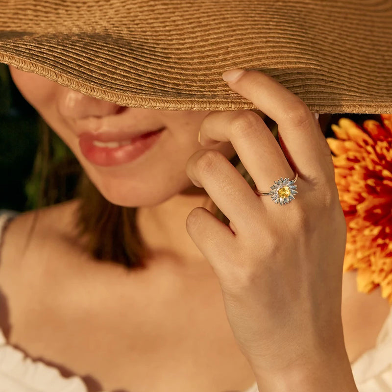 Woman wearing a straw hat and a ring with a yellow gemstone, holding an orange flower.