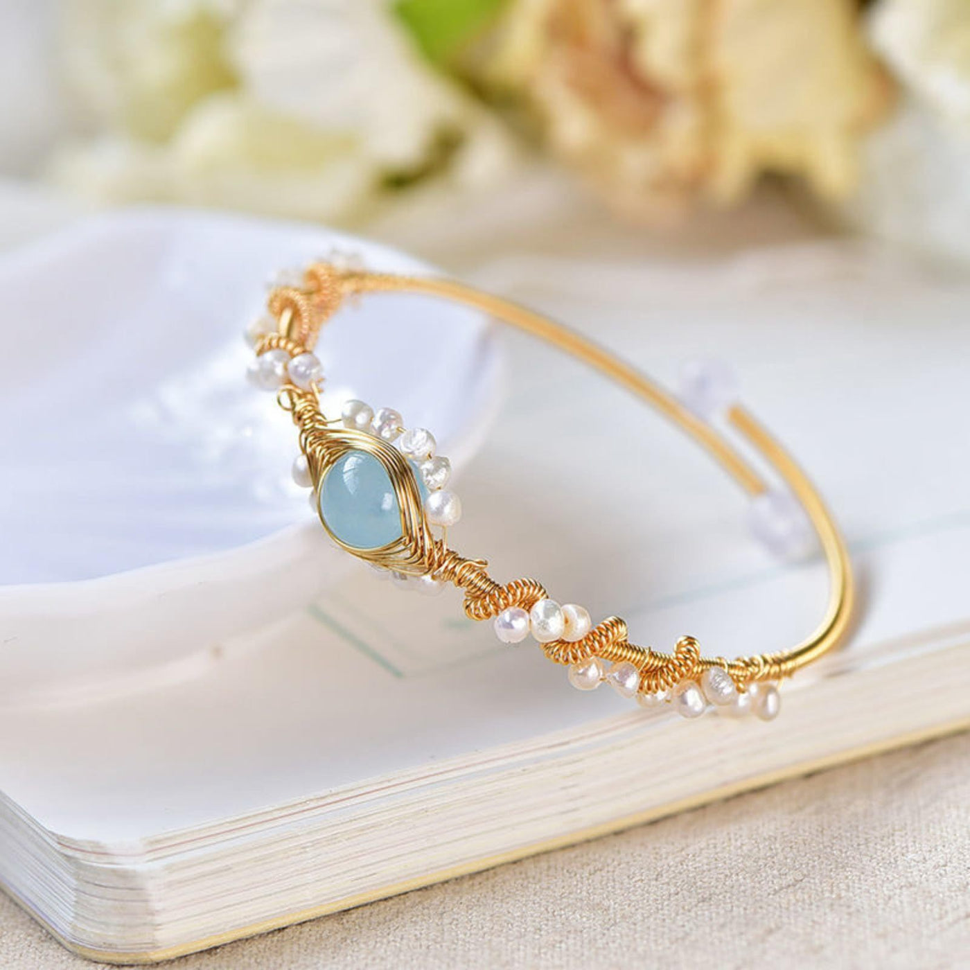 Gold bracelet with pearls and a blue gemstone on a white surface with flowers in the background