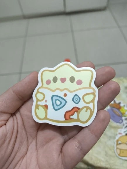 Hand holding a colorful cartoon-style sticker on a tiled floor background