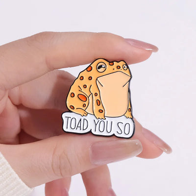 Enamel pin with a cartoon toad and 'TOAD YOU SO' text held by a hand on a white background