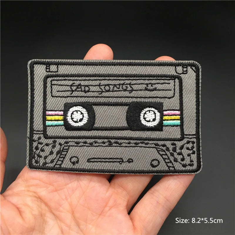 Embroidered patch of a cassette tape with 'Sad Songs' text held in a hand against a dark background.