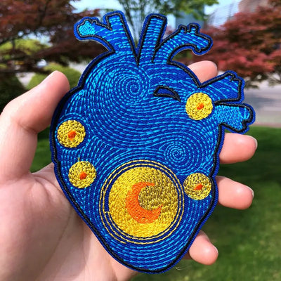 Blue and yellow embroidered heart-shaped patch held in a hand with a blurred outdoor background
