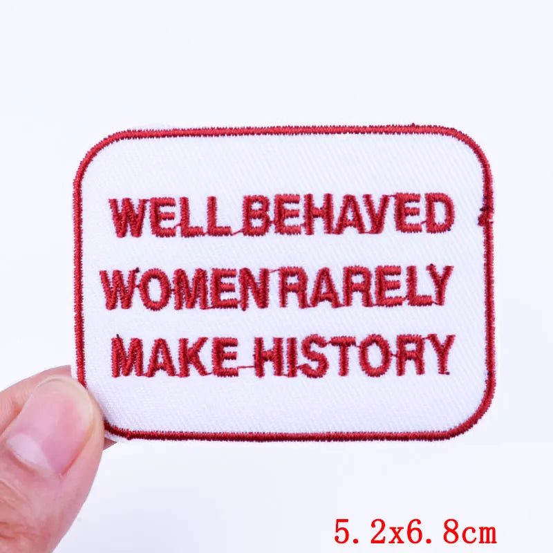 Embroidered patch with text 'WELLBEHAVED WOMEN RARELY MAKE HISTORY' held by a hand on a white background.