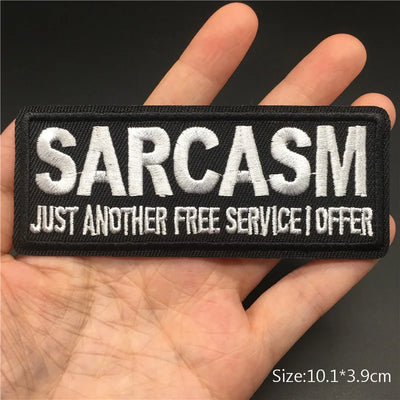 Patch with 'SARCASM JUST ANOTHER FREE SERVICE I OFFER' text held in a hand against a black background