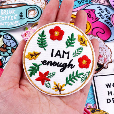 Hand holding a patch with 'I AM enough' text surrounded by flowers and leaves, with other patches in the background.