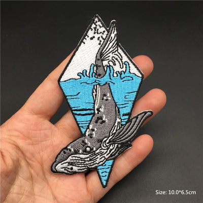Embroidered patch of a whale and iceberg held in a hand against a dark background