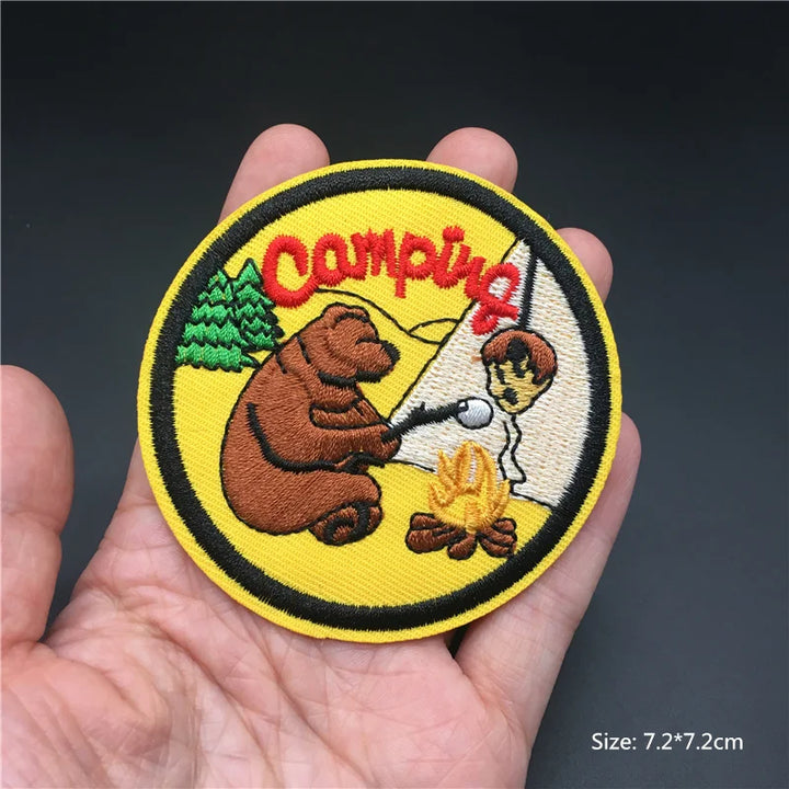 Camping-themed patch with a bear and campfire design held in a hand against a dark background.