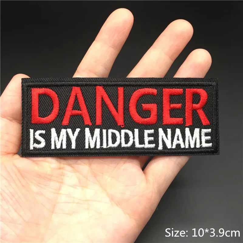 Patch with 'DANGER IS MY MIDDLE NAME' text held in a hand against a dark background