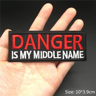Patch with 'DANGER IS MY MIDDLE NAME' text held in a hand against a dark background