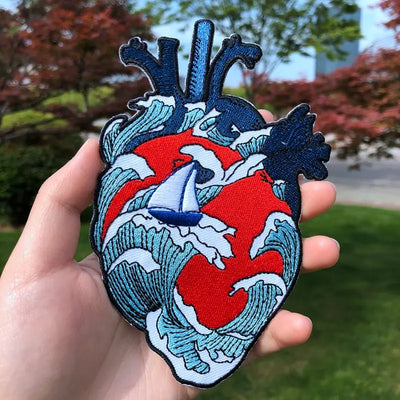Heart-shaped patch with wave design and sailboat held in a hand outdoors.