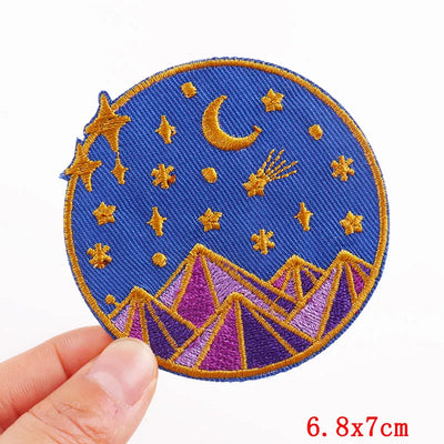 Embroidered patch with stars, moon, and mountains held by a hand on a white background