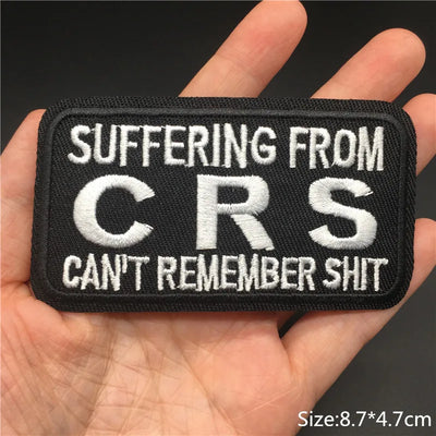 Patch with text 'SUFFERING FROM CRS CAN'T REMEMBER SHIT' held in a hand against a dark background