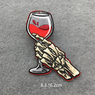 Embroidered patch of a skeleton hand holding a glass of red wine on a gray background.