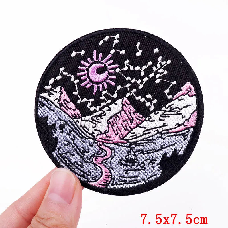 Circular embroidered patch with pink and white design held by a hand, on a white background.