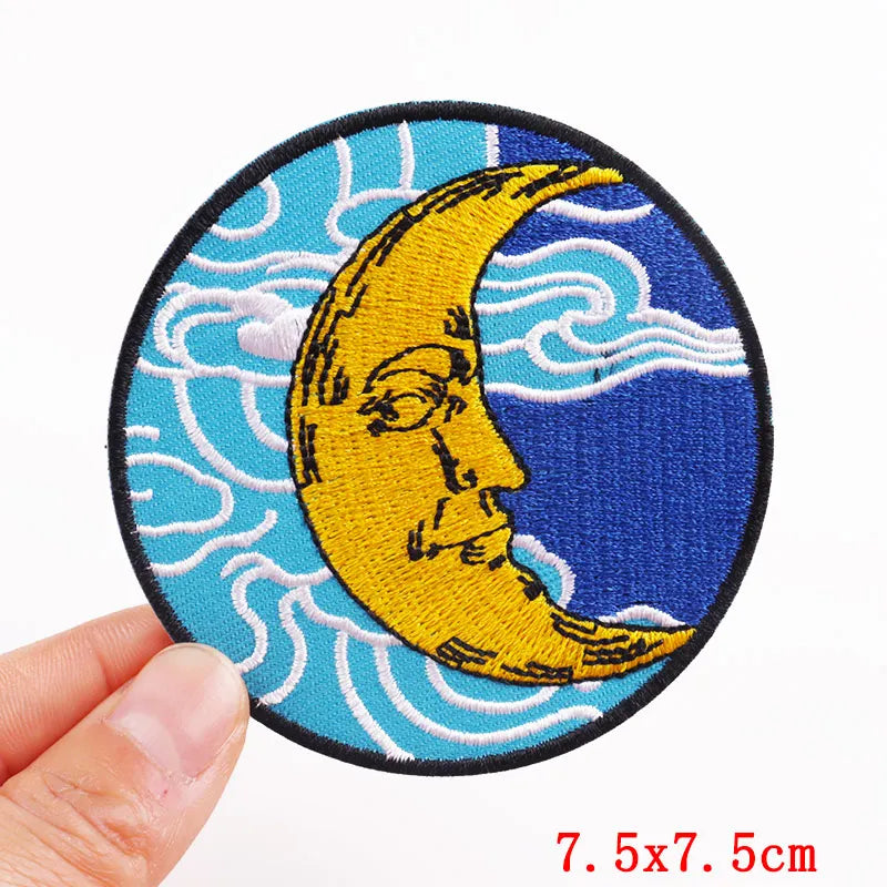 Circular patch with a yellow crescent moon and face design on a blue background, held by a hand.