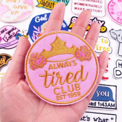 Pink patch with 'Always Tired Club' text held in a hand, surrounded by other patches.