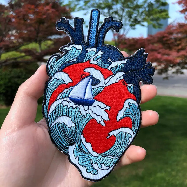 Heart-shaped patch with wave design and sailboat held in a hand outdoors.