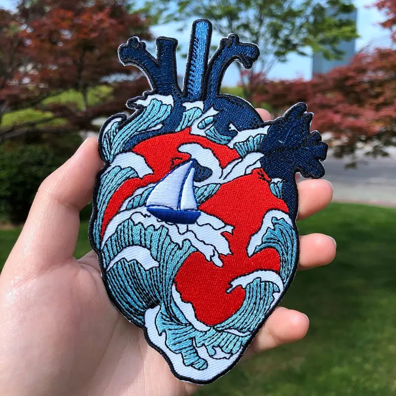 Heart-shaped patch with wave design and sailboat held in a hand outdoors.