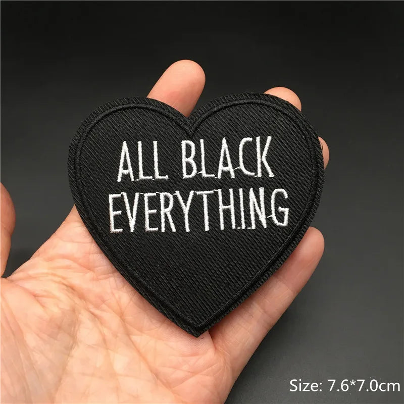 Heart-shaped patch with 'ALL BLACK EVERYTHING' text held in a hand against a dark background