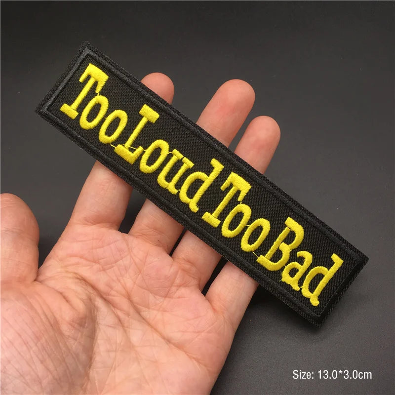 Embroidered patch with 'Too Loud Too Bad' text held in a hand against a dark background