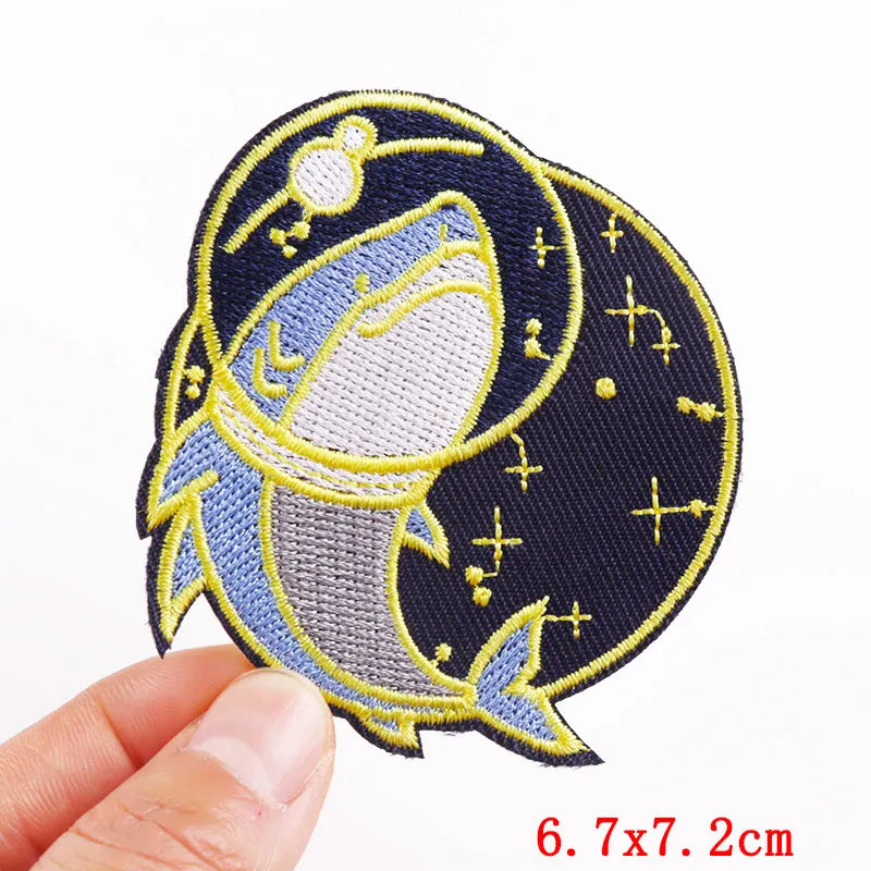 Embroidered patch of a shark with starry background held by a hand, size 6.7x7.2cm.