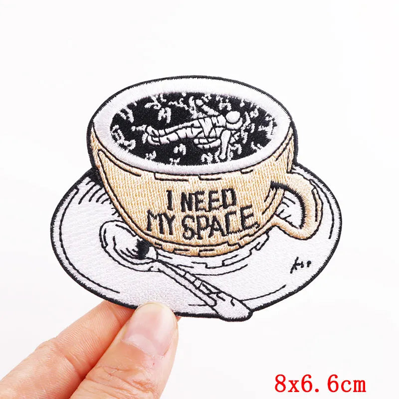 Embroidered patch of a coffee cup with 'I need my space' text, held by a hand.