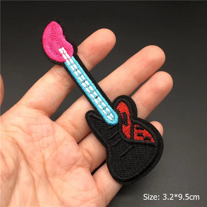 Embroidered patch of a guitar with pink flame design held in a hand against a dark background.