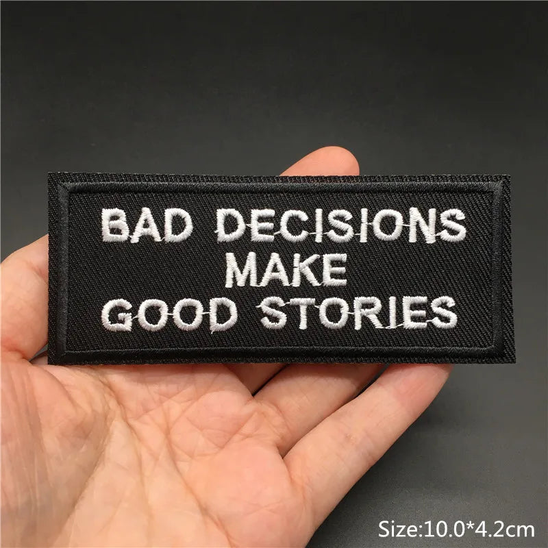Black patch with white text 'BAD DECISIONS MAKE GOOD STORIES' held in a hand against a dark background.