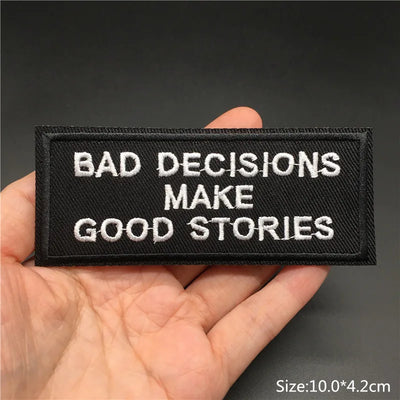 Black patch with white text 'BAD DECISIONS MAKE GOOD STORIES' held in a hand against a dark background.