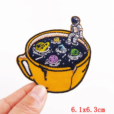 Embroidered patch of a cup with a space-themed design held by a hand, on a white background.