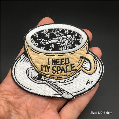 Embroidered patch of a coffee cup with 'I Need My Space' text, held in a hand against a dark background.