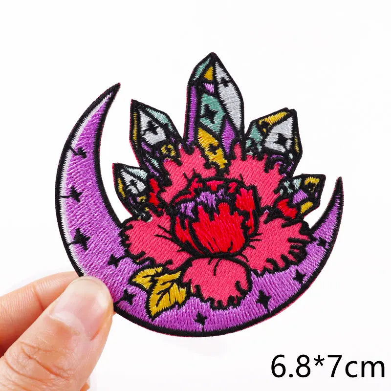 Colorful embroidered patch with a pink flower and purple crescent moon held by a hand on a white background.