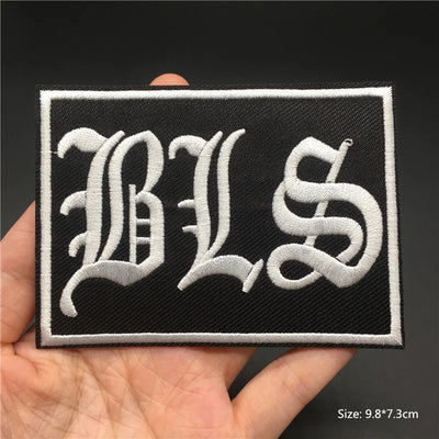 Black patch with white 'BLS' letters held in a hand against a dark background