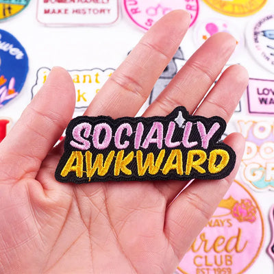 Hand holding a patch with 'SOCIALLY AWKWARD' text against a background of colorful patches.